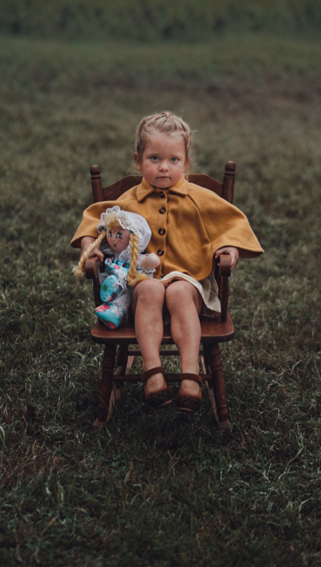 Little girl in mustard cape sitting in rocking chair with doll